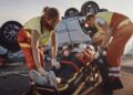 Two paramedics render emergency care to an injured person stabilized on a stretcher. A rolled-over vehicle lies behind them.