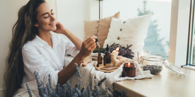 A young woman adding essential oils to a home diffuser. She has a lavender plant, a candle, and pillows near her.