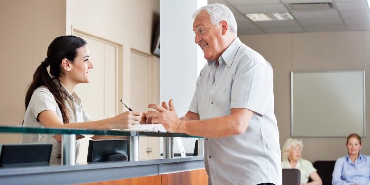 An older adult man talking to a receptionist at a medical clinic while two other patients sit in chairs in the background.