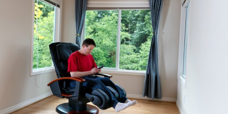 A homeowner using his massage chair at home. He's wearing comfortable clothing and using the remote to start his session.