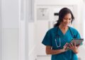 A female healthcare worker wears turquoise scrubs, a stethoscope, and a hospital badge. She is holding and using a tablet.