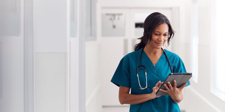 A female healthcare worker wears turquoise scrubs, a stethoscope, and a hospital badge. She is holding and using a tablet.