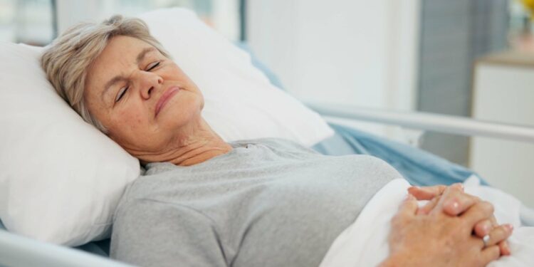 A woman lies in a hospital bed with closed eyes and a gentle smile. Her hands are on her abdomen, and her head is slightly raised.