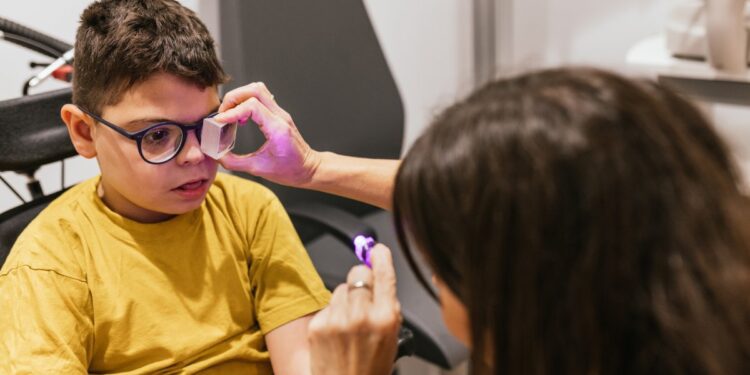 An optometrist uses a purple light to check a child's eyes in the exam room. The child sits in a wheelchair.