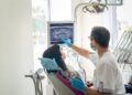 A licensed dentist pointing at an X-ray for a patient. The patient is sitting in the cleaning chair in a pink shirt.