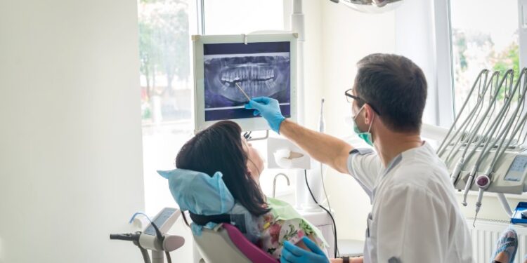 A licensed dentist pointing at an X-ray for a patient. The patient is sitting in the cleaning chair in a pink shirt.