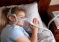 A woman wearing a blue shirt and a CPAP machine is sleeping on her side on a floral-patterned pillow.